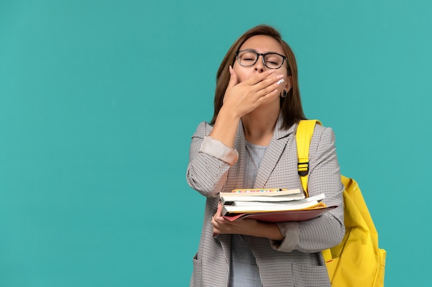 Vista frontale della studentessa in zaino giallo giacca grigia che tiene i libri che sbadigliano sulla parete blu