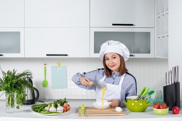 Vista frontale dello chef felice e verdure fresche con attrezzatura da cucina e miscelazione dell'uovo in una ciotola bianca nella cucina bianca