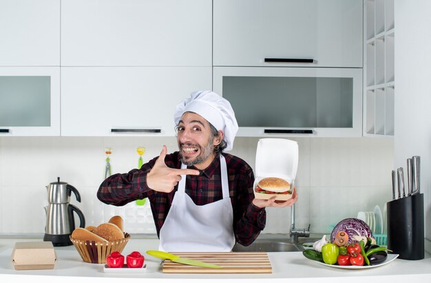 Vista frontale dello chef maschio che indica l'hamburger in mano in cucina