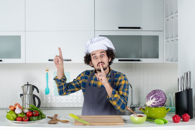 Vista frontale dello chef maschio con verdure fresche e cucina con utensili da cucina e gesto di silenzio rivolto verso l'alto nella cucina bianca