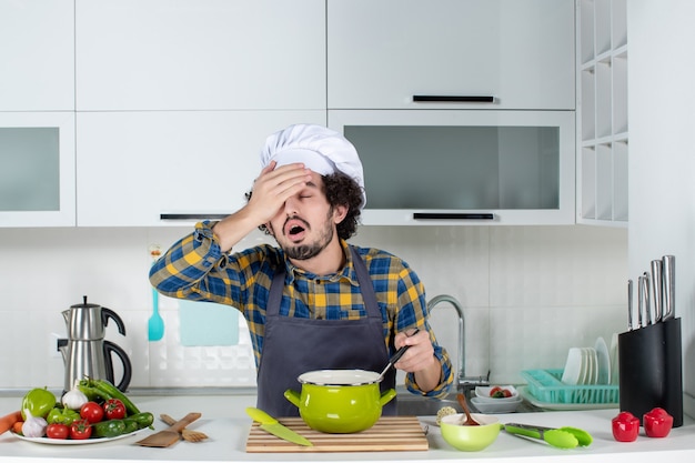 Vista frontale dello chef maschio con verdure fresche e tenendo il cucchiaio nel pasto sentendosi esausto nella cucina bianca