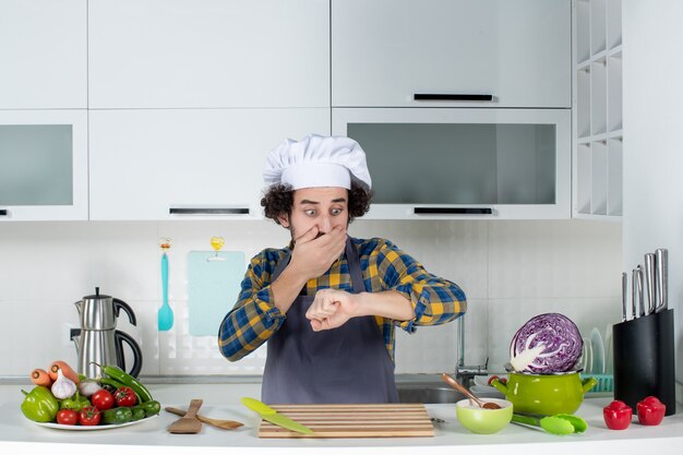 Vista frontale dello chef maschio scioccato con verdure fresche e cucina con utensili da cucina e controlla il suo tempo nella cucina bianca white