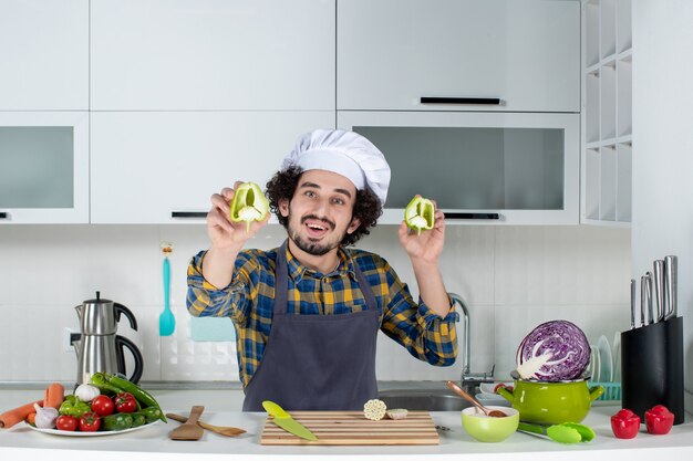 Vista frontale dello chef maschio sorridente con verdure fresche e cucina con utensili da cucina e mostra peperoni verdi tagliati nella cucina bianca