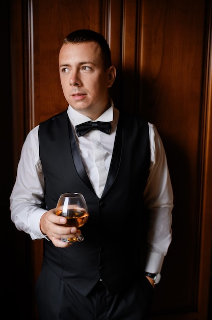 Vista frontale dello sposo pensieroso che indossa un elegante abito nero e una camicia bianca con cravatta a farfalla con in mano un bicchiere pieno di bevanda
