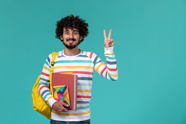 Vista frontale dello studente maschio che indossa lo zaino giallo che tiene file e quaderni sulla parete blu