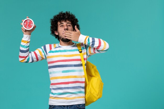 Vista frontale dello studente maschio che indossa lo zaino giallo che tiene gli orologi che sbadigliano sulla parete azzurra