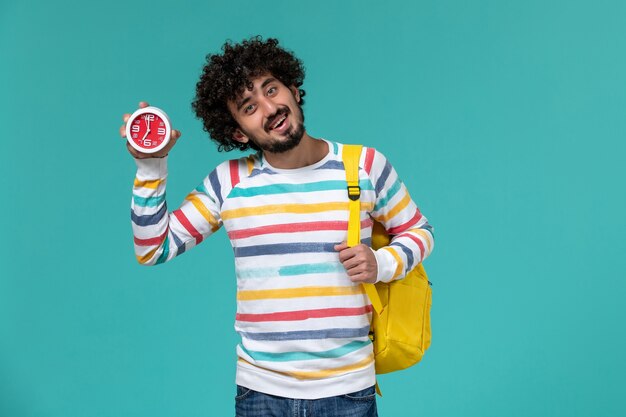 Vista frontale dello studente maschio che indossa lo zaino giallo che tiene gli orologi sulla parete blu-chiaro