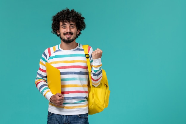 Vista frontale dello studente maschio in camicia a righe che indossa lo zaino giallo che tiene i file sorridenti sulla parete blu