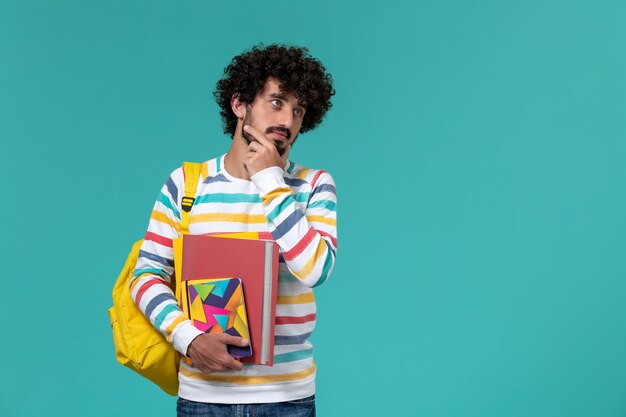 Vista frontale dello studente maschio in camicia a righe colorate che indossa lo zaino giallo che tiene file e quaderni pensando sulla parete blu