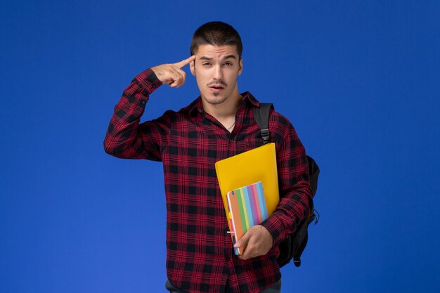 Vista frontale dello studente maschio in camicia a scacchi rossa con lo zaino che tiene file e quaderni sulla parete blu