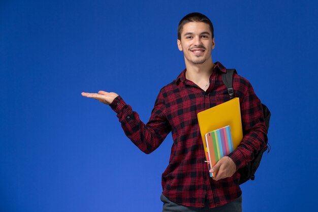 Vista frontale dello studente maschio in camicia a scacchi rossa con lo zaino che tiene file e quaderni sulla parete blu