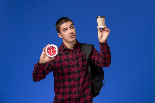 Vista frontale dello studente maschio in camicia a scacchi rossa con lo zaino che tiene gli orologi e il caffè sulla parete blu