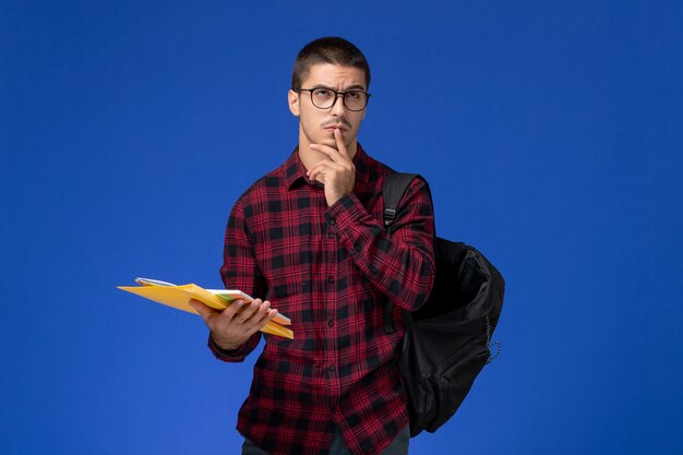 Vista frontale dello studente maschio in camicia a scacchi rossa con lo zaino che tiene i file e il quaderno pensando sulla parete azzurra