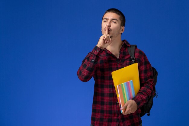 Vista frontale dello studente maschio in camicia a scacchi rossa con lo zaino che tiene i file e quaderni che mostrano il segno di silenzio sulla parete blu