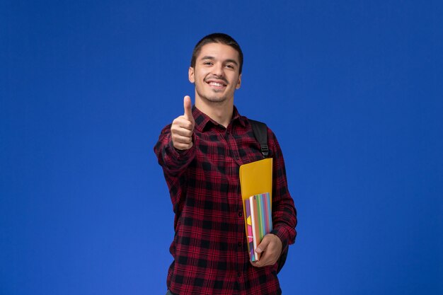Vista frontale dello studente maschio in camicia a scacchi rossa con lo zaino che tiene i file e quaderni sorridente sulla parete blu