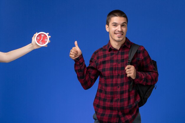 Vista frontale dello studente maschio in camicia a scacchi rossa con lo zaino sorridente sulla parete blu