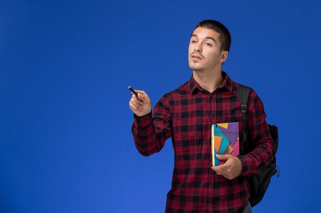 Vista frontale dello studente maschio in camicia a scacchi rossa con zaino che tiene il quaderno sulla parete azzurra
