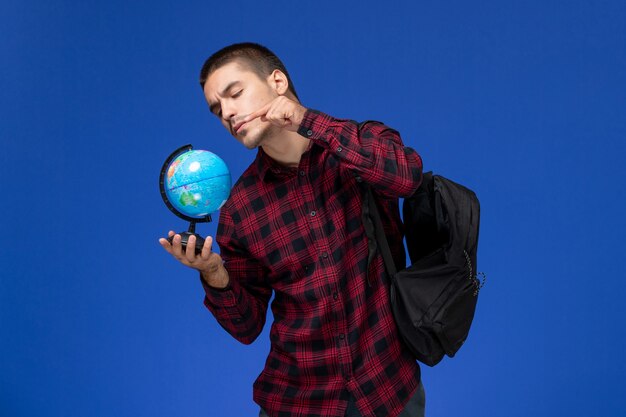 Vista frontale dello studente maschio in camicia a scacchi rossa con zaino che tiene piccolo globo sulla parete azzurra