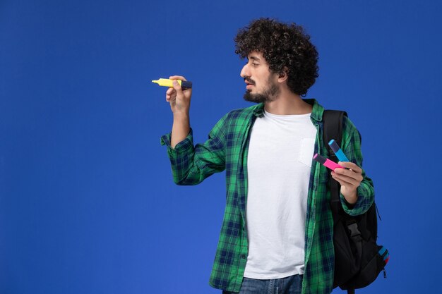 Vista frontale dello studente maschio in camicia a scacchi verde con zaino nero che tiene i pennarelli sulla parete blu