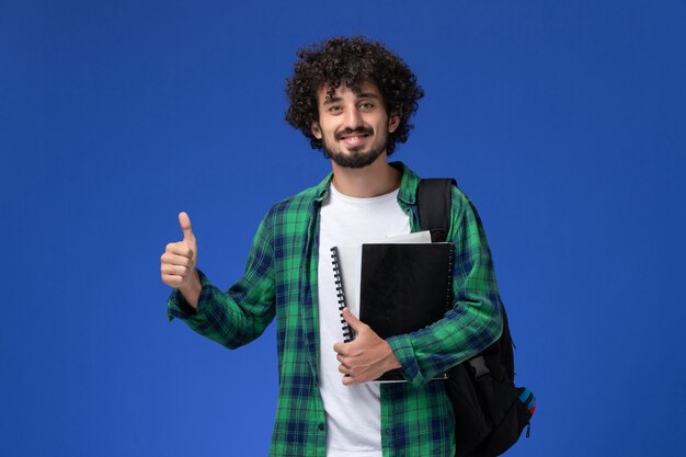 Vista frontale dello studente maschio in camicia a scacchi verde con zaino nero che tiene i quaderni sorridente sulla parete blu