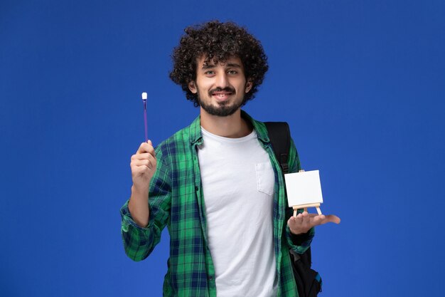 Vista frontale dello studente maschio in camicia a scacchi verde con zaino nero che tiene nappa e cavalletto sulla parete blu