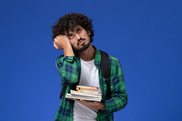 Vista frontale dello studente maschio in camicia a scacchi verde con zaino nero che tiene quaderni e file pensando sulla parete blu