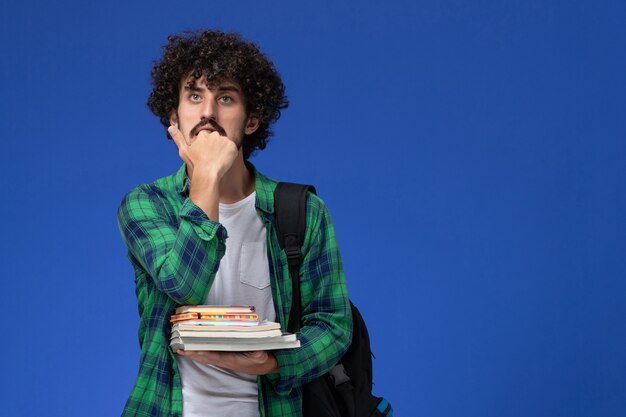 Vista frontale dello studente maschio in camicia a scacchi verde con zaino nero che tiene quaderni e file sulla parete blu chiaro