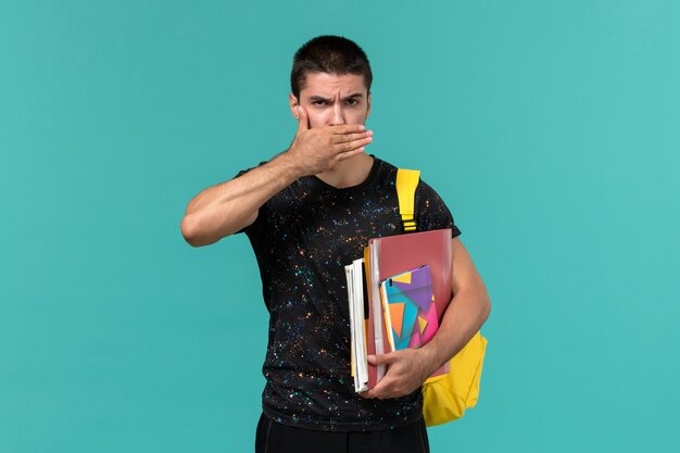 Vista frontale dello studente maschio in maglietta scura che indossa lo zaino giallo che tiene il quaderno e file che chiudono la bocca sulla parete blu