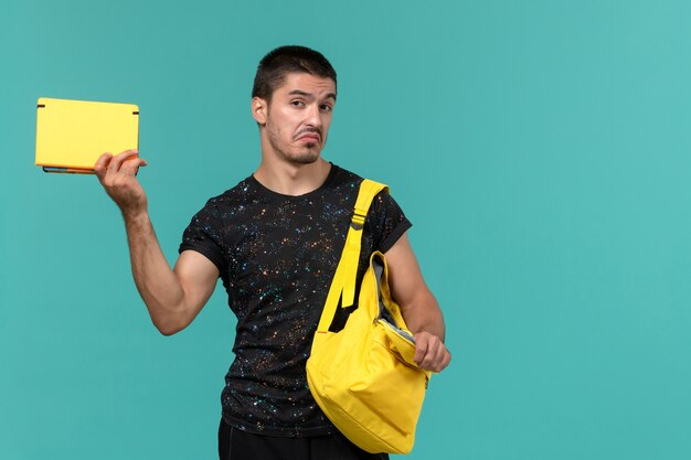 Vista frontale dello studente maschio in t-shirt scura zaino giallo che tiene il quaderno sulla parete blu chiaro