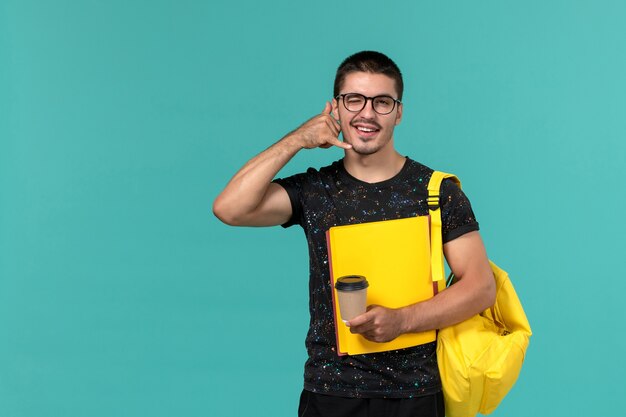 Vista frontale dello studente maschio in zaino giallo t-shirt scura che tiene diversi file e caffè sulla parete blu