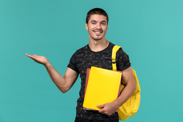 Vista frontale dello studente maschio in zaino giallo t-shirt scura che tiene diversi file sulla parete blu chiaro