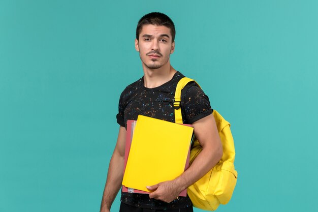 Vista frontale dello studente maschio in zaino giallo t-shirt scura che tiene diversi file sulla parete blu chiaro