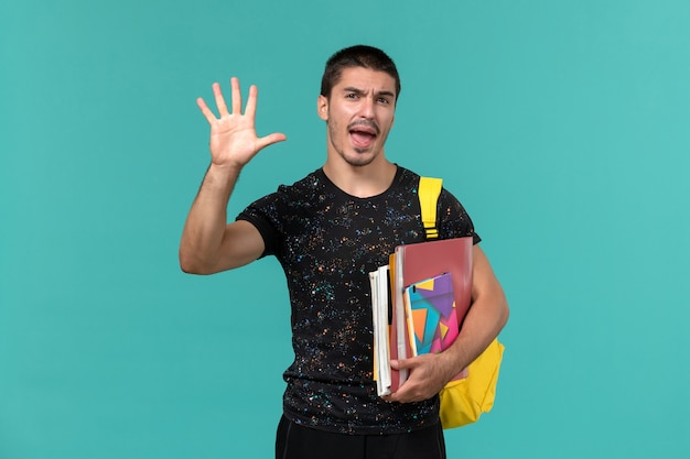 Vista frontale dello studente maschio in zaino giallo t-shirt scura che tiene quaderno e file sulla parete blu