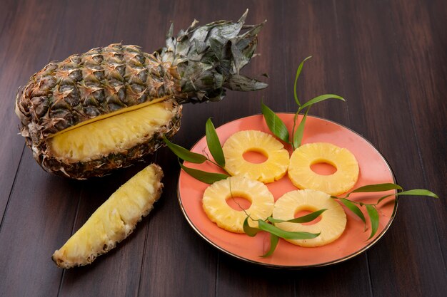 Vista frontale di ananas con un pezzo tagliato da frutta intera con fette di ananas in lamiera su superficie di legno