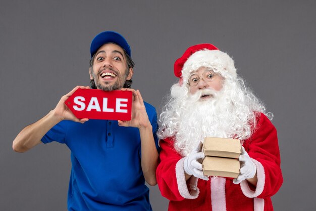 Vista frontale di Babbo Natale con corriere maschio che tiene banner di vendita e pacchetti di cibo sul muro grigio