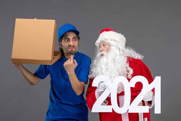 Vista frontale di Babbo Natale con corriere maschio che tiene i sacchetti della spesa e la scatola di cibo sul muro grigio