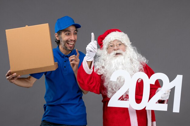 Vista frontale di Babbo Natale con corriere maschio che tiene la scatola di cibo e sul muro grigio