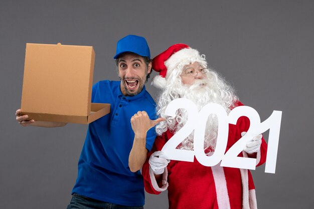 Vista frontale di Babbo Natale con corriere maschio che tiene la scatola di cibo e sul muro grigio