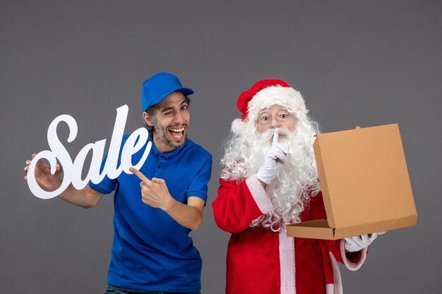 Vista frontale di Babbo Natale con corriere maschio che tiene la scrittura di vendita e la scatola di cibo sul muro grigio