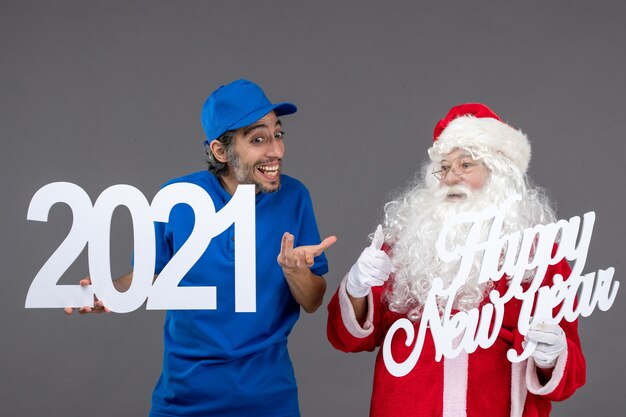 Vista frontale di Babbo Natale con corriere maschio che tiene schede di felice anno nuovo e 2021 sul muro grigio