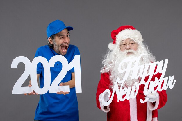Vista frontale di Babbo Natale con corriere maschio che tiene schede di felice anno nuovo e 2021 sul muro grigio