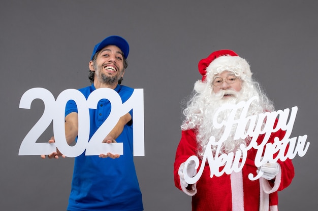 Vista frontale di Babbo Natale con corriere maschio che tiene schede di felice anno nuovo e 2021 sul muro grigio