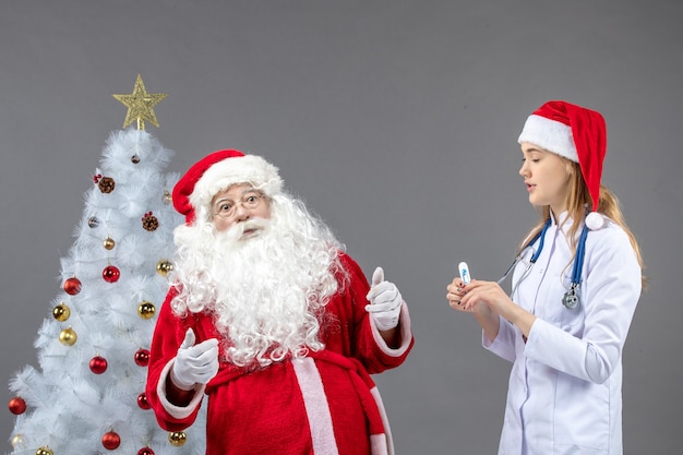 Vista frontale di Babbo Natale con dottoressa che tiene il dispositivo di misurazione della temperatura sul muro grigio