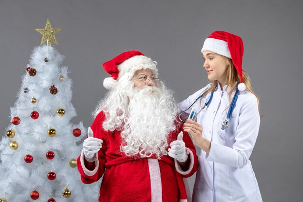 Vista frontale di Babbo Natale con dottoressa che tiene il dispositivo di misurazione della temperatura sul muro grigio