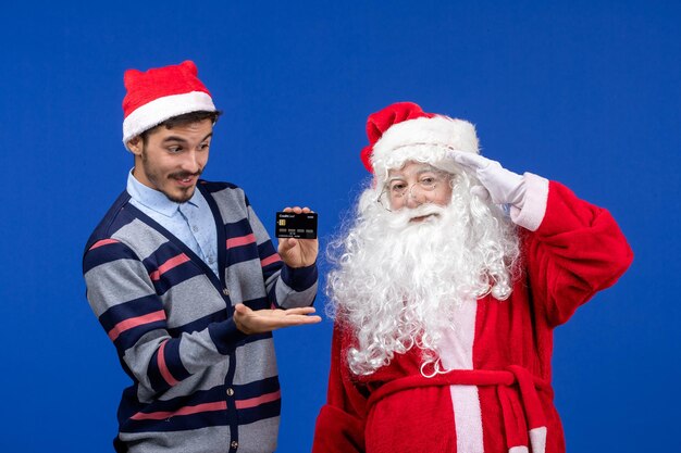 Vista frontale di babbo natale con il giovane che tiene la carta bancaria sul muro blu