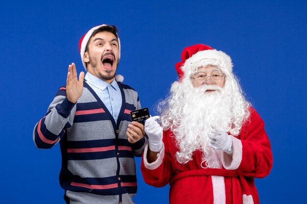 Vista frontale di babbo natale con il giovane che tiene la carta bancaria sul muro blu