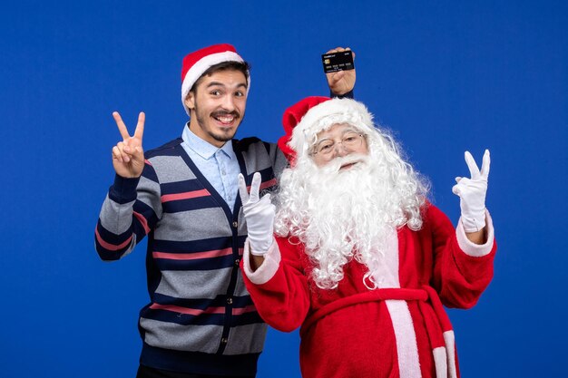 Vista frontale di babbo natale con il giovane che tiene la carta bancaria sul muro blu