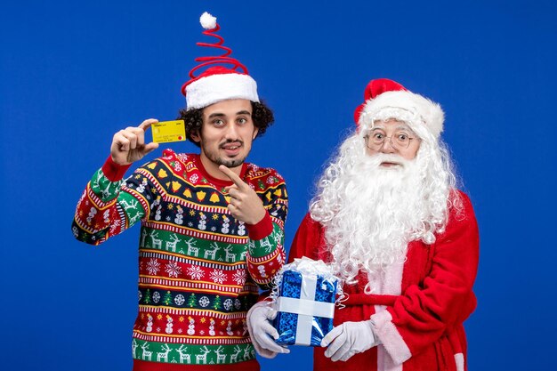 Vista frontale di babbo natale con il giovane che tiene la carta di credito e presente sul muro blu