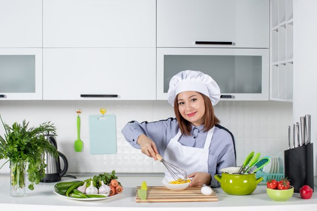 Vista frontale di chef fiducioso e verdure fresche con attrezzatura da cucina e mescolando l'uovo in una ciotola bianca nella cucina bianca