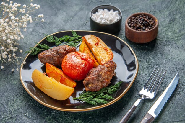 Vista frontale di deliziose cotolette di carne al forno con patate e pomodori su un piatto nero set di posate fiori bianchi spezie su sfondo verde nero colori misti